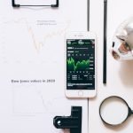 Vista superior de ferramentas de análise financeira, incluindo um celular, prancheta, lupa e cofrezinho em uma mesa branca.