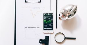 Vista superior de ferramentas de análise financeira, incluindo um celular, prancheta, lupa e cofrezinho em uma mesa branca.