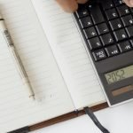 Uma mão usando uma calculadora com documentos financeiros e um caderno, retratando uma configuração de escritório para análise financeira.