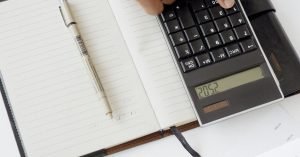 Uma mão usando uma calculadora com documentos financeiros e um caderno, retratando uma configuração de escritório para análise financeira.